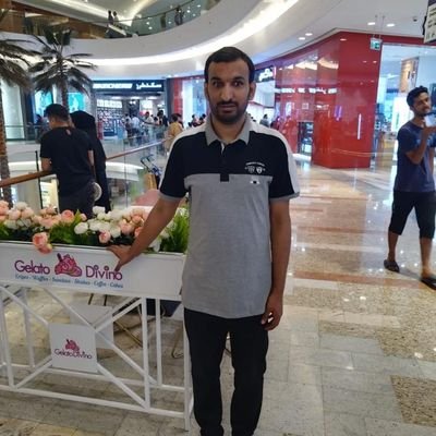 Hammad, a man with medium brown skin and short dark hair, stands in a brightly lit shopping mall beside a white Gelato DiVino counter decorated with pink and white flowers. He faces the camera with a neutral expression, wearing a black-and-gray short-sleeved polo shirt, black pants, and black shoes. The shiny tiled floor, multiple levels of the mall with curved balconies and glass railings, and several shoppers and storefronts in the background create a busy indoor atmosphere.